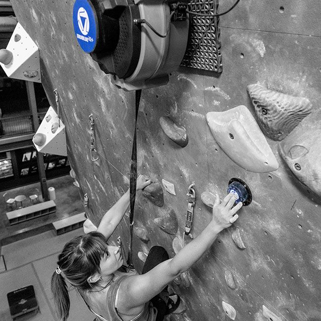 woman climbing using a TRUBLUE Auto Belay Device on an indoor climbing wall in black and white with blue highlights on auto belay and chalk bag