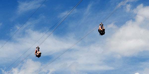 two people on twin zip lines with a partially cloudy blue sky as background